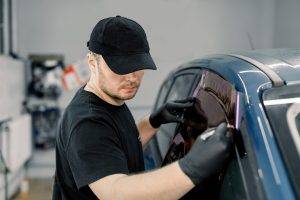 Closeup image of a handsome car mechanic worker, wearing black uniform, attaching tinting foil to car window in specialized service station.