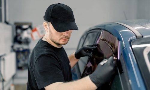 Closeup image of a handsome car mechanic worker, wearing black uniform, attaching tinting foil to car window in specialized service station.